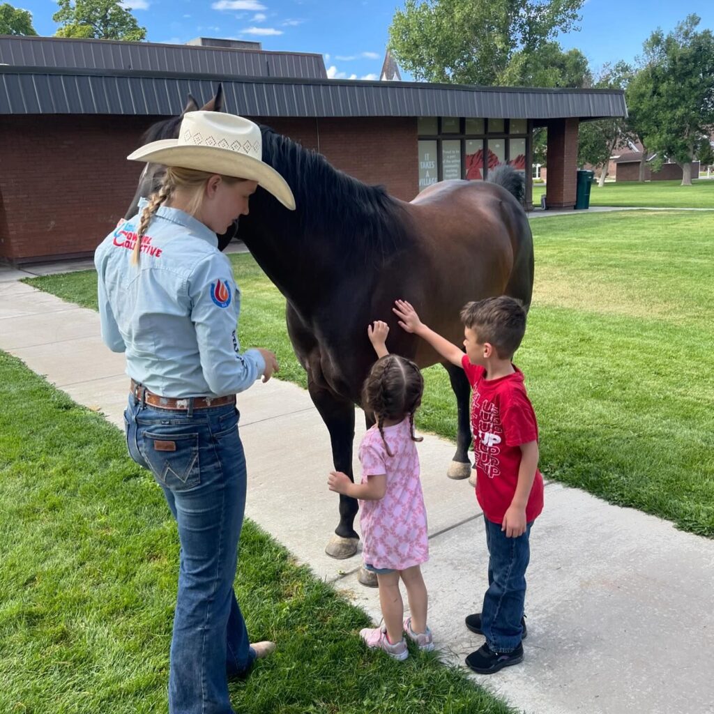 A young woman in a cowboy hat encourages young children to pet her pony