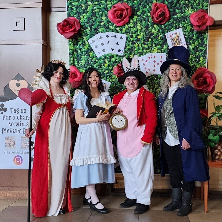 Four people are dressed as characters from "Alice's Adventures in Wonderland". They are standing in front of a beautiful backdrop that features red roses and playing cards against a green background.