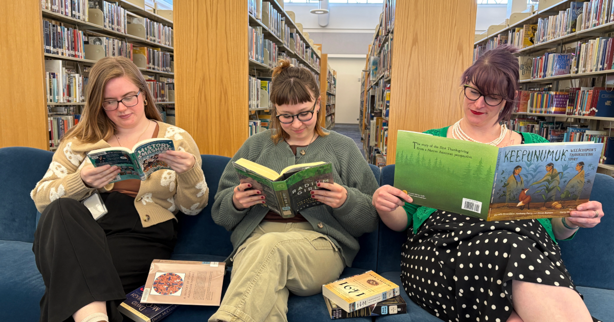 Three women read books on a comfy couch in a library.