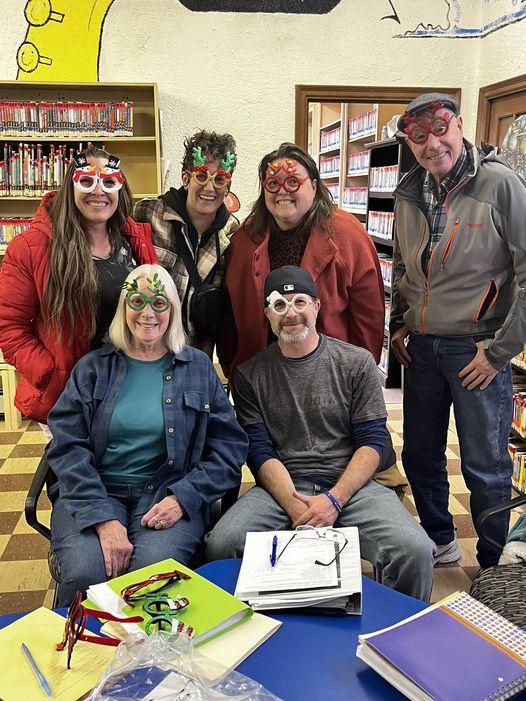 A group of six happy people are wearing wacky Christmas glasses inside a library.